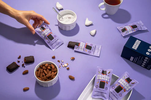 Hand reaching for Krack’d Snacks Dark Chocolate Aloha Caramel bar among keto-friendly snack ingredients.