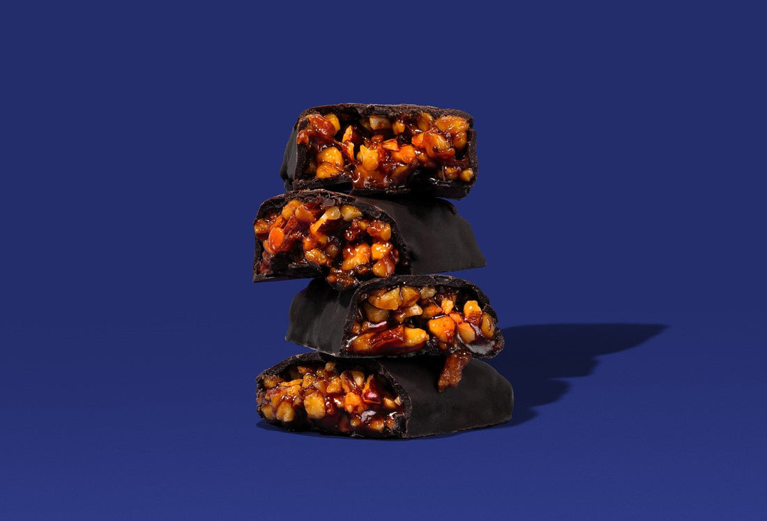 Four dark chocolate-covered nut bars from Krack’d Snacks are stacked on top of each other against the white background.