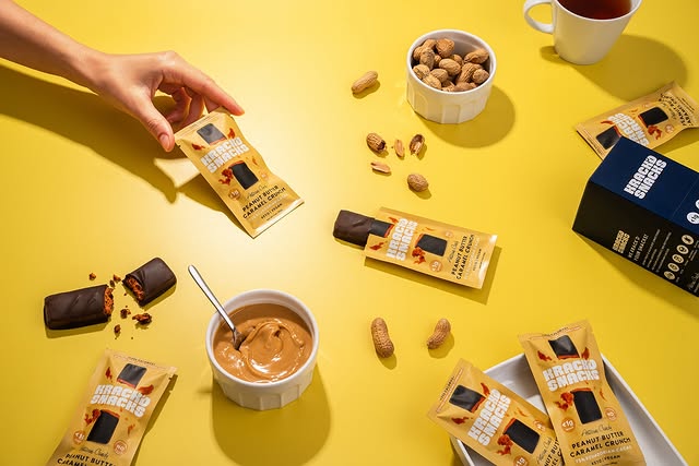 A hand picks up a "Krack'd Snacks Dark Chocolate Peanut Butter Caramel Crunch" bar surrounded by various keto-friendly snacks.